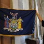 Die Flagge von New York besteht aus dem Wappen des Bundesstaats auf blauem Hintergrund. Es zeigt zwei Schildhalterinnen:<br />Links: Die Göttin Libertas mit einer Phrygischen Mütze auf ihrem Stab. Ihr linker Fuß steht auf einer Krone als Symbol für die Freiheit von Großbritannien.<br />Rechts: Die Göttin Justitia mit Waage und Schwert.
