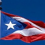 Ein Willkommensschild habe ich nicht - deshalb geht's gleich zur Flagge. Die Flagge Puerto Ricos besteht aus fünf gleich großen, horizontalen Streifen in den Farben rot und weiß.<br /><br />Auf der linken Seite werden die Streifen durch ein blaues, gleichschenkliges Dreieck überdeckt. In ihm ist ein weißer, fünfzackiger Stern dargestellt.<br /><br />Die Farbe Rot symbolisiert das Blut der mutigen Männer, Weiß den Sieg und den Frieden, und die Farbe Blau den Himmel und das Meer, das die Insel Puerto Rico umgibt. Der Stern ist Symbol für die Einheit der Nation, während das Dreieck die drei Gewalten des Staates darstellt. Sie wurde 1894 von Francisco Gonzalo Marín entworfen.<br /><br />Die Flagge ähnelt stark der Flagge Kubas, wobei die Farben blau und rot vertauscht und das Seitenverhältnis auf 2:3 verkürzt sind. 1895 wurde von der Sektion Puerto Rico der Partido Revolucionario Cubano (Revolutionären Kubanischen Partei) in New York dieser Entwurf als offizielle Fahne der Befreiungsbewegung Puerto Ricos festgelegt.<br /><br />Erst seit 1952 darf die Fahne öffentlich rechts neben der Flagge der USA gezeigt werden, während sie bis dahin als Fahne der illegalen Befreiungsbewegung Puerto Ricos galt. Bei Sportveranstaltungen wie Olympischen Spielen war bis dahin ein an die Gouverneursflagge angelehnter weißer Banner mit Emblem und „Puerto Rico“-Schriftzug in Gebrauch.<br />