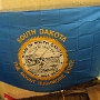 Die Flagge des US-Bundesstaats South Dakota wurde im Jahr 1963 angenommen.<br /><br />Die Flagge von South Dakota wurde auf der Basis früherer Entwürfe eingeführt, die ursprünglich das Motto „The Sunshine State" trugen. Im Jahr 1992 wurde das Motto zu „The Mount Rushmore State" geändert.<br /><br />Das Siegel selbst zeigt eine Landschaft mit Symbolen für Industrie, Landwirtschaft und Verkehr. Am oberen Rand des Siegels befindet sich ein Spruchband mit dem Motto des Bundesstaats:<br />„Under God the people rule.“